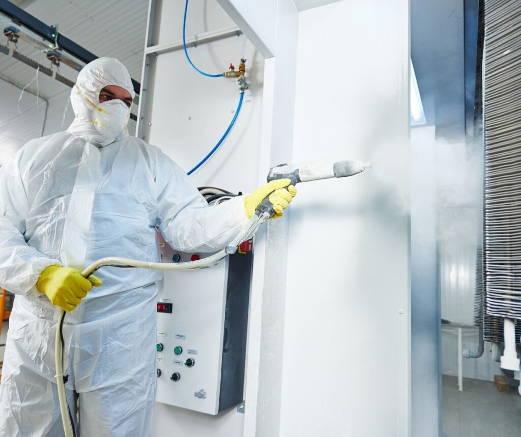 industrial metal coating. Worker man in protective suit with gas mask spraying powder to steel finished parts in painting chamber