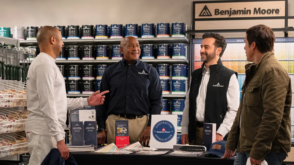 Benjamin Moore 2026 Contractor Appreciation Month 1 Four men talking at a Benjamin Moore paint store counter during a contractor appreciation event, with shelves of paint cans and color samples in the background.